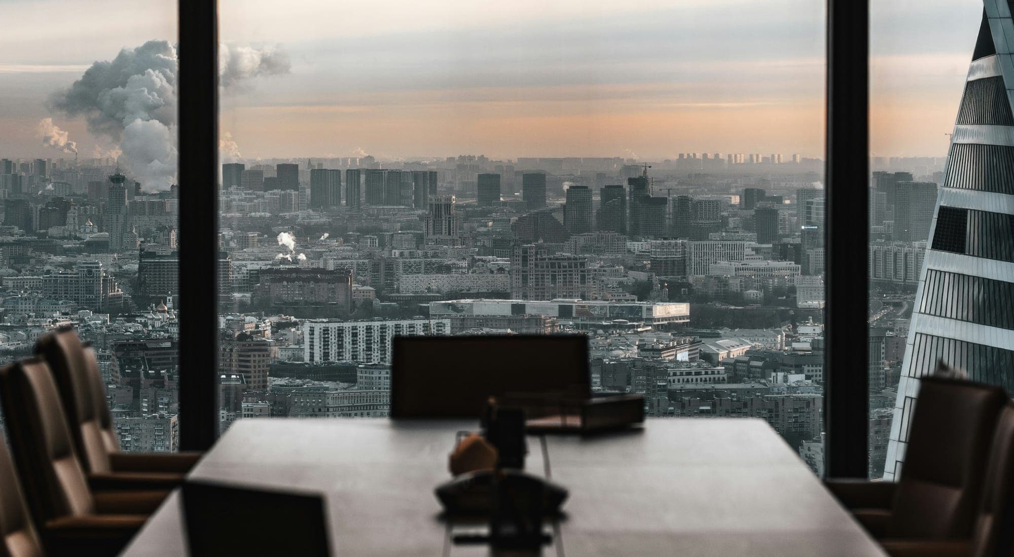 An empty boardroom table before a window overlooking a city at dusk